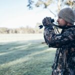 Adult male in camouflage outfit using binoculars in an open field, showcasing outdoor exploration and observation.